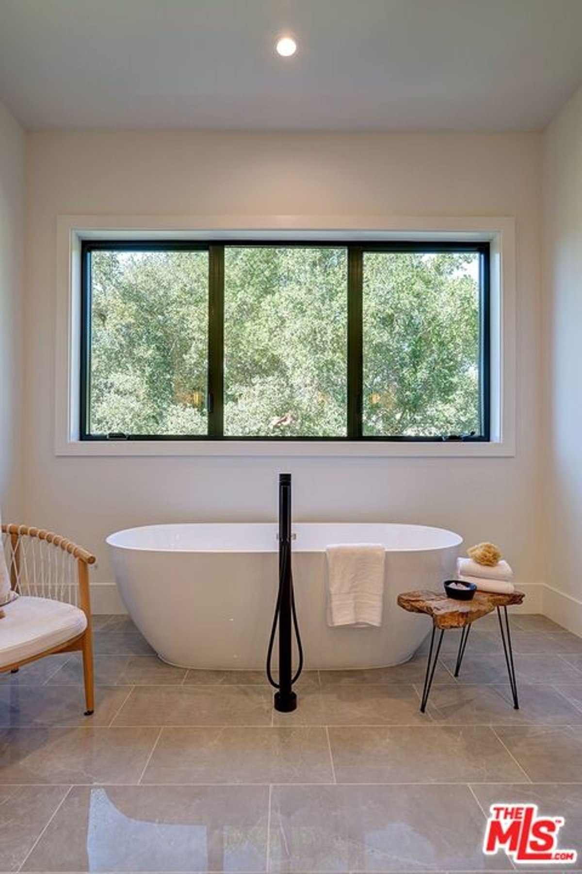 Freestanding white bathtub is positioned in front of a wide window, paired with a rustic wooden side table.