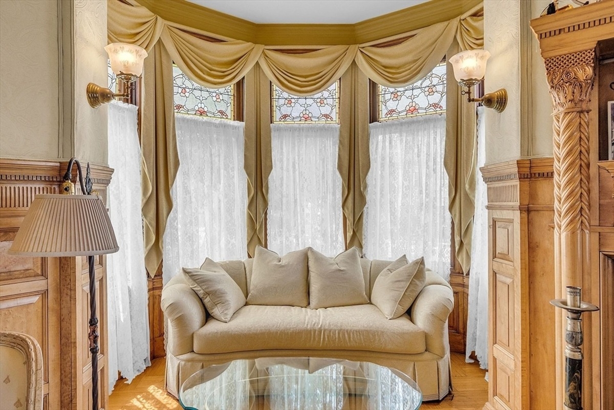 Comfortable cream-colored sofa is framed by large windows with stained glass and flowing curtains.