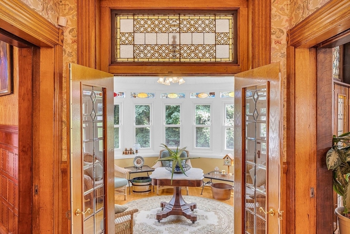 Sunroom with bright white walls is surrounded by large windows with colorful stained glass accents and wicker furniture.