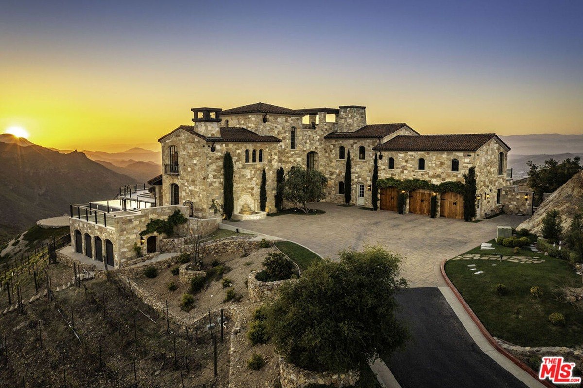 Stone-clad mansion overlooking the mountainous landscape at sunset with a serene driveway.