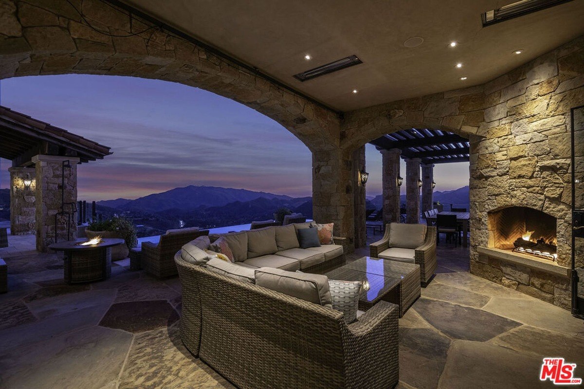 Covered outdoor lounge with stone arches, comfortable seating, and a fireplace overlooking the sunset-lit hills.