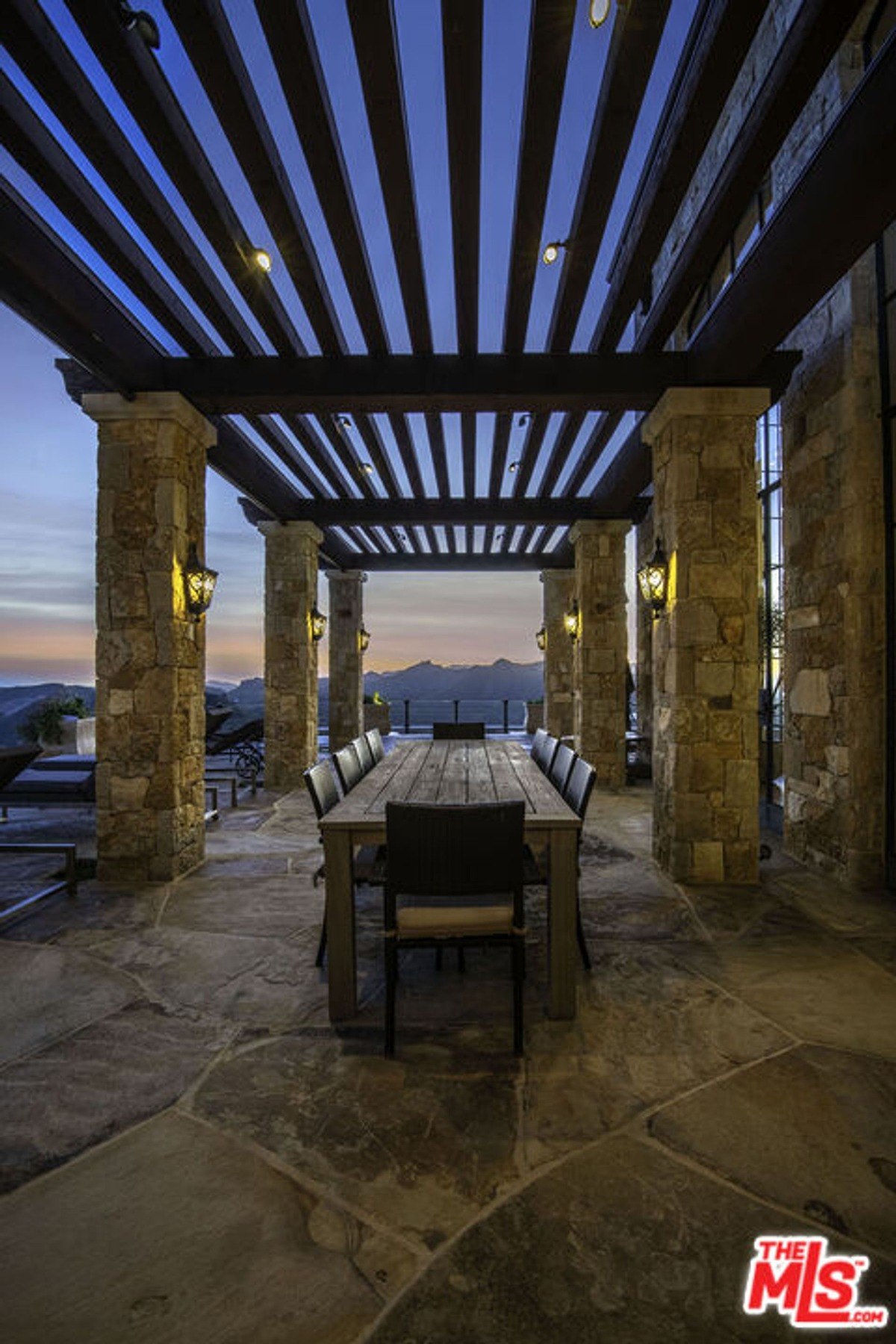 Rustic dining area on a stone-paved terrace framed by stone columns and overhead pergola beams.