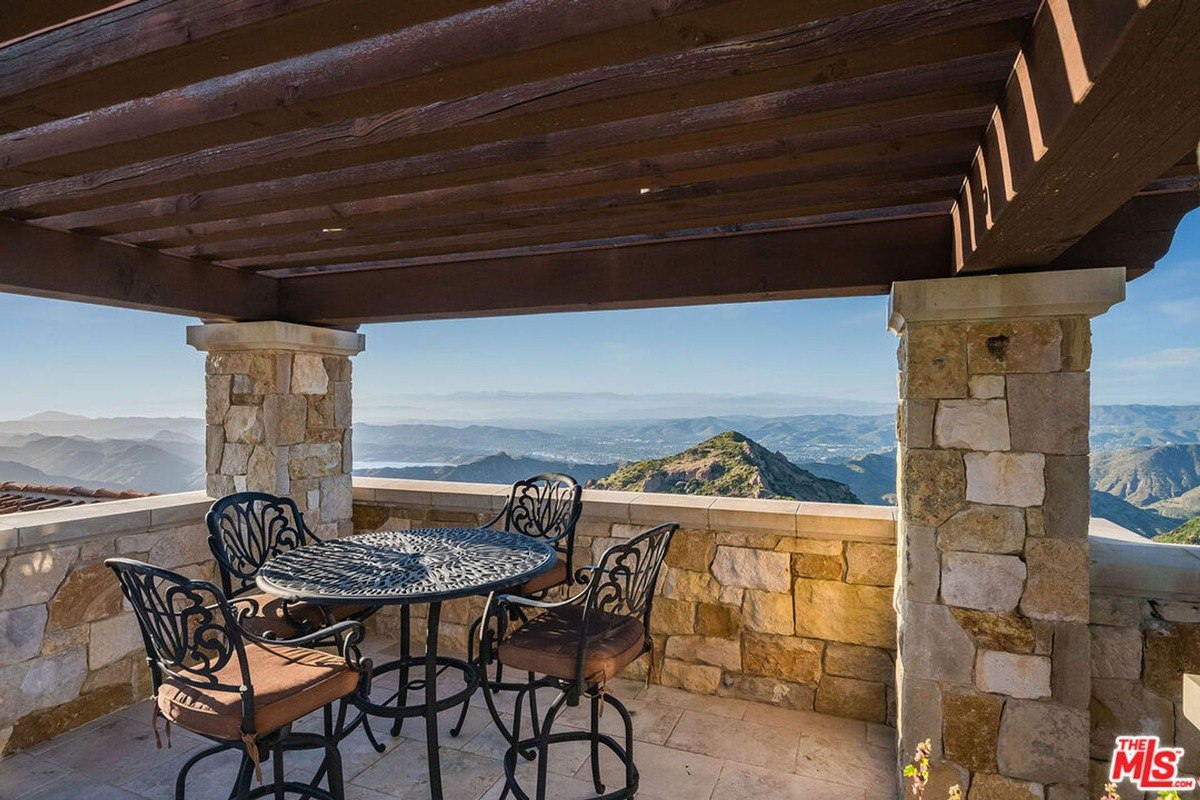 Stone patio has a metal table and chairs under a wooden pergola, overlooking a mountain range.