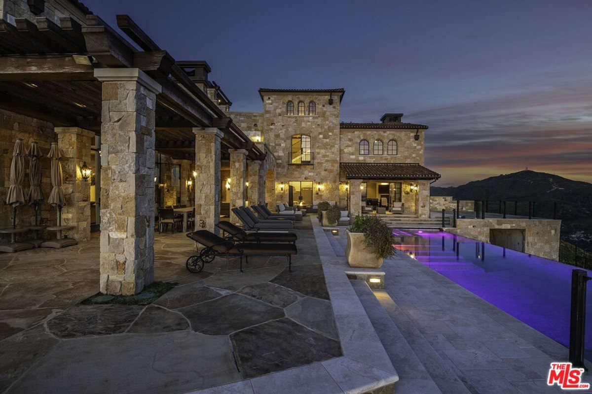 Modern stone house has a large pool and patio area with lounge chairs at dusk.