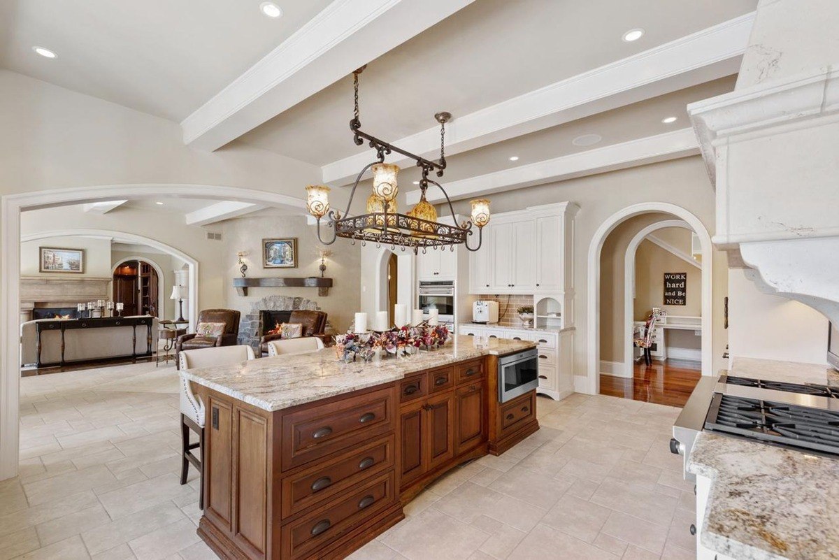 Warm neutral tones and a coffered ceiling create a welcoming atmosphere in this spacious kitchen, featuring a large island, white cabinetry, and views into the living area.