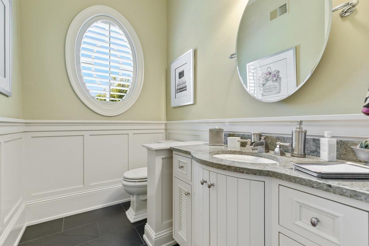 Powder room with a circular window, white wainscoting, and a granite-topped vanity offers a bright and elegant touch.