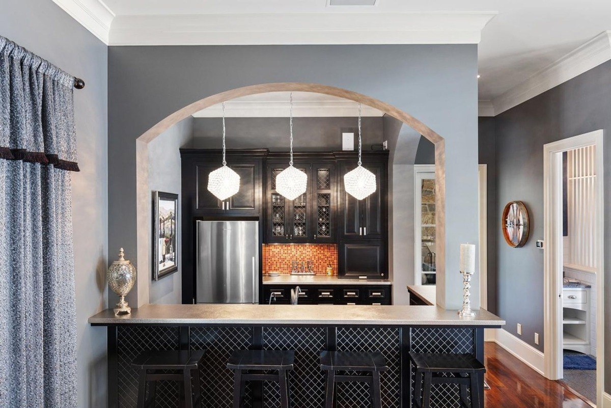 Stylish bar area with a sleek counter, pendant lights, and an arched opening into a well-equipped kitchen offers a sophisticated space for entertaining.