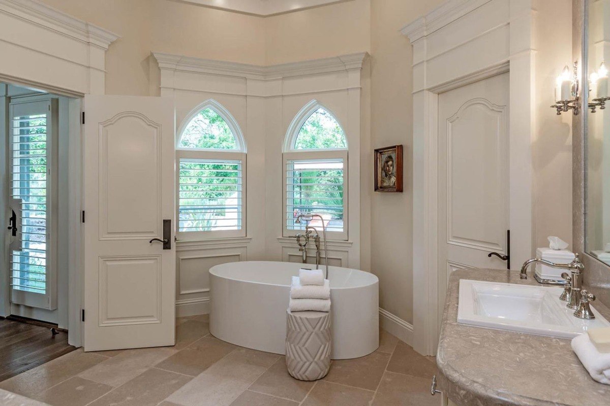 Luxurious freestanding soaking tub set beneath arched windows with shutters.