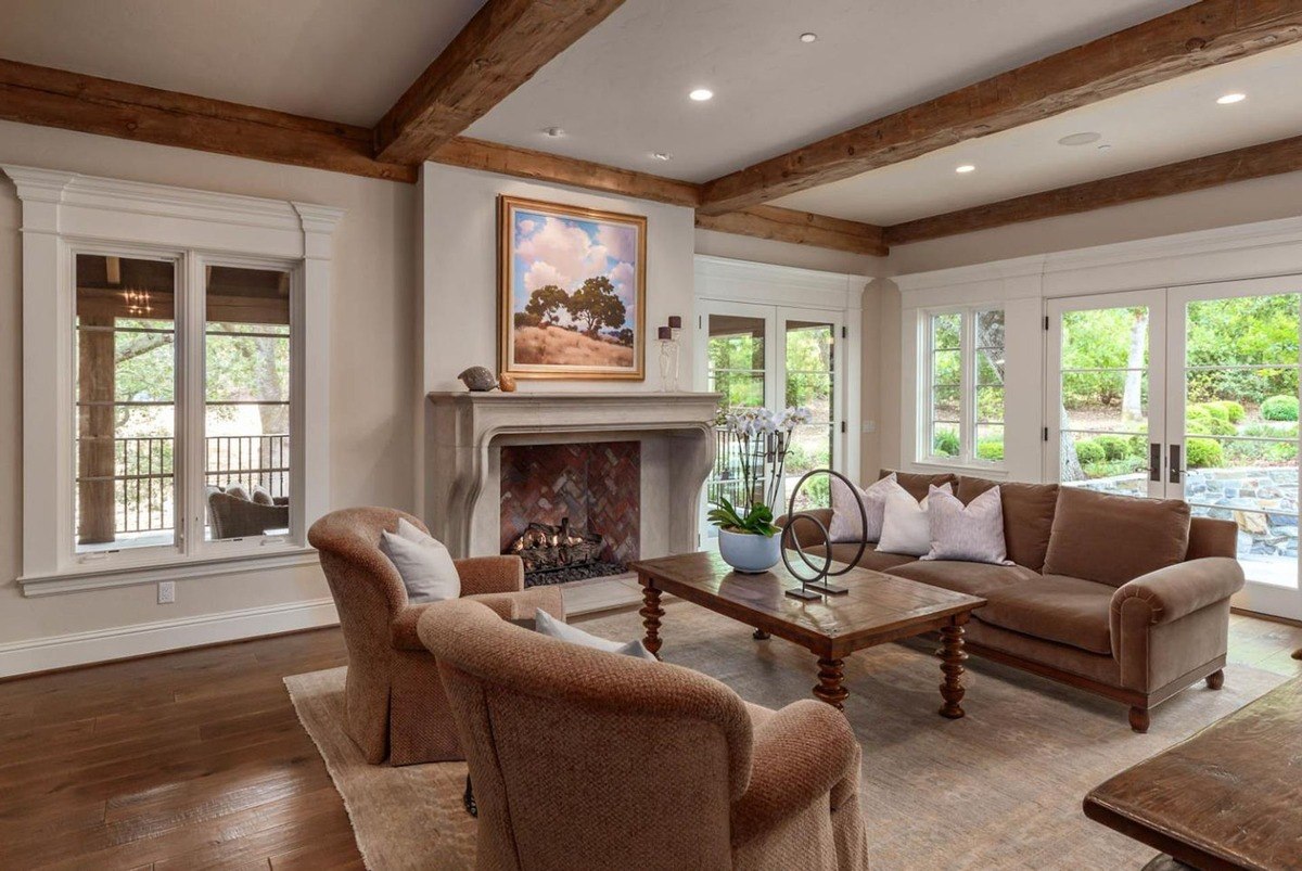 Living room with beamed ceiling, stone fireplace, and large windows.