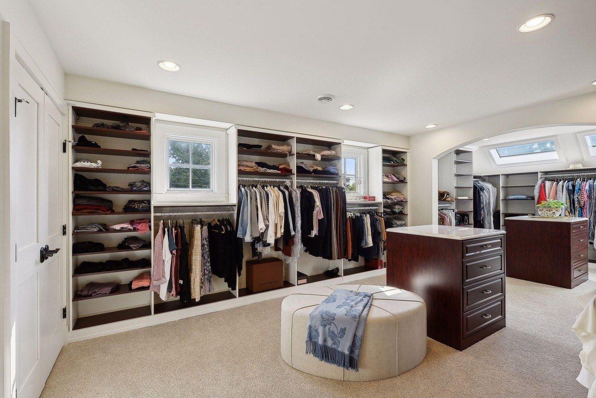 Large walk-in closet with ample shelving, hanging rods, and a comfortable ottoman.