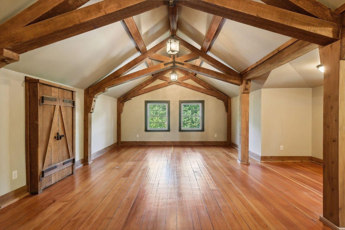 Large room with exposed wooden beams, hardwood floors, and two windows.