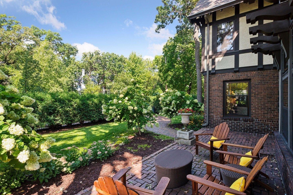 Brick patio with wooden chairs and a fire pit, adjacent to a house and lush green yard.