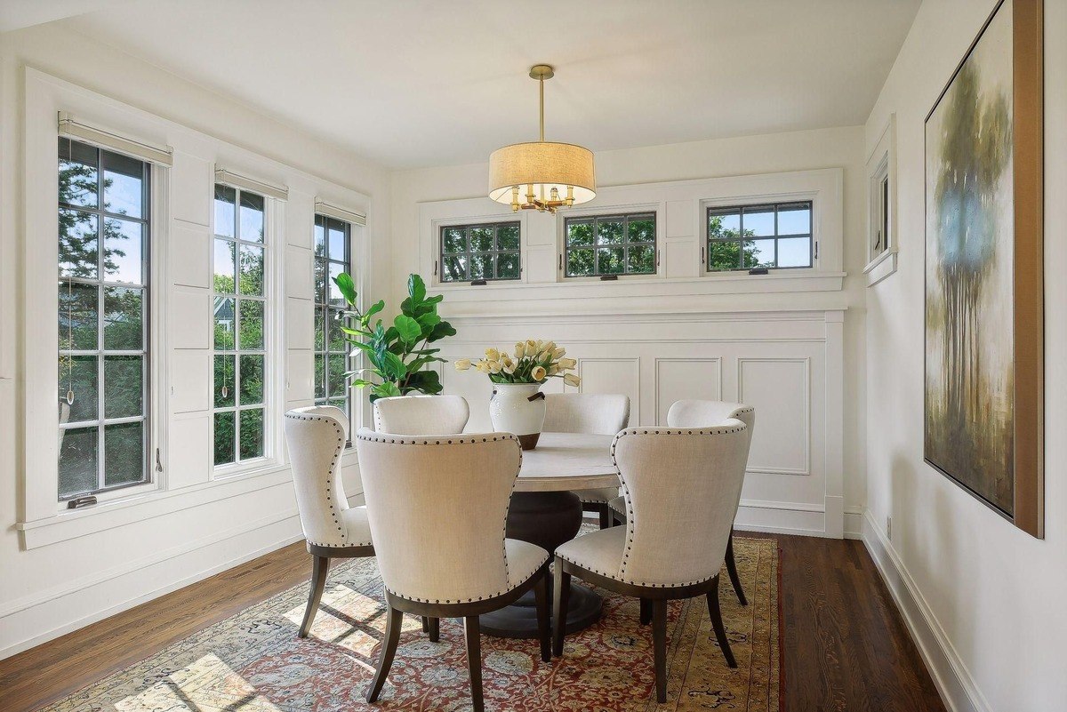 Dining room with white chairs surrounding a round table, hardwood floors, and large windows.