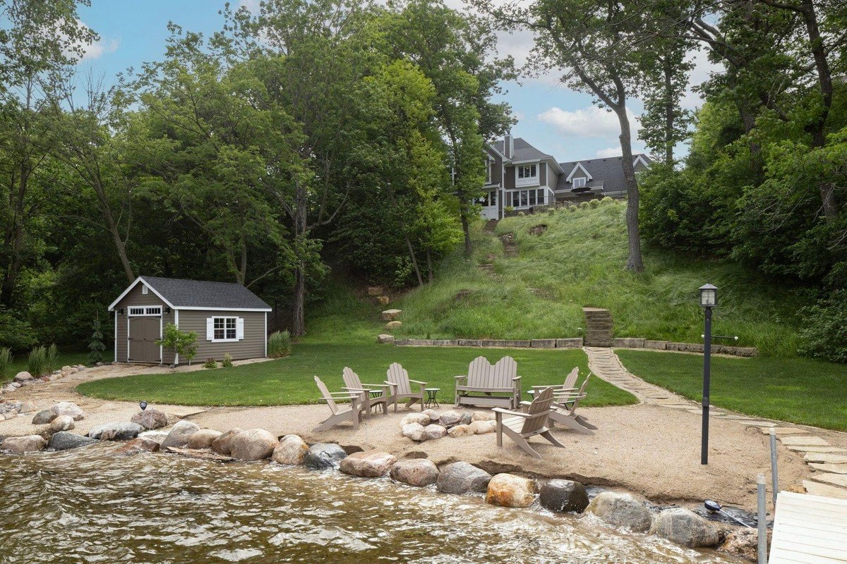 Lakeside retreat features a fire pit surrounded by Adirondack chairs, a path leading to a shed, and a grand home nestled among the trees.