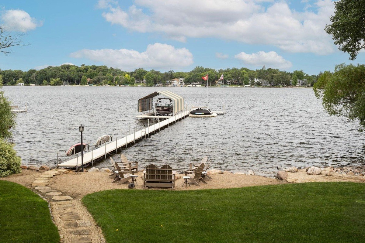 Sandy beach with a path leading to a dock extends into a calm lake, offering a picturesque view under a bright blue sky.