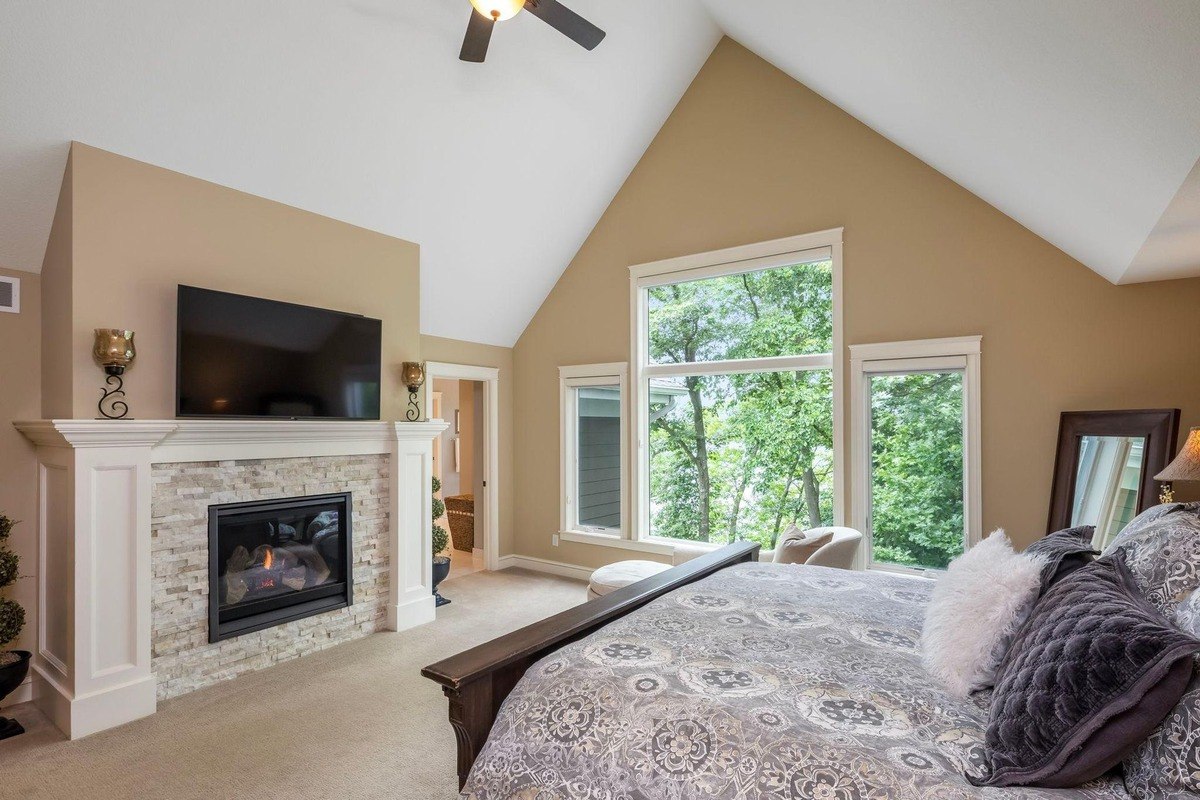 Vaulted-ceiling primary bedroom with a fireplace and large windows facing greenery.