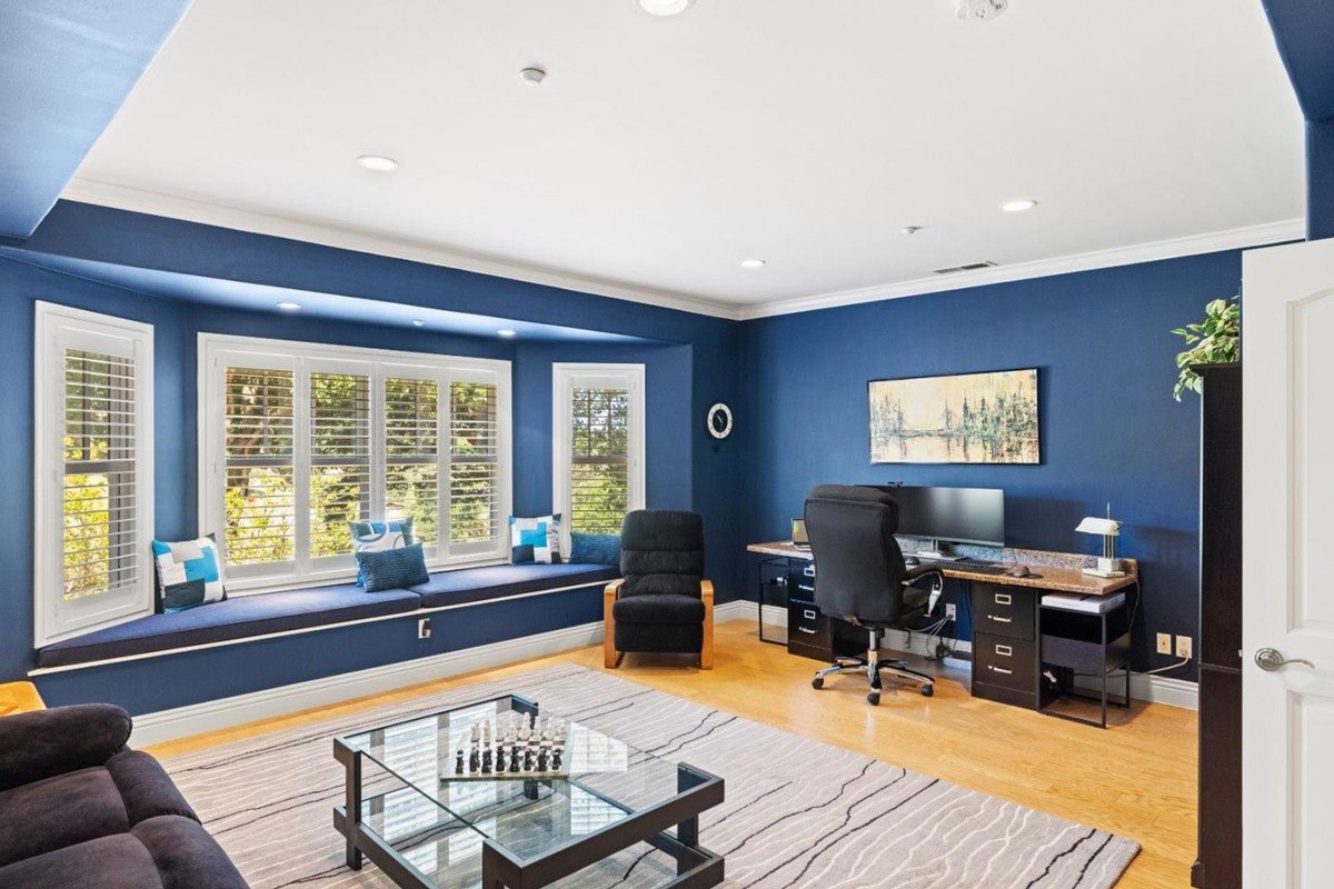 Versatile room with a bay window seating area, a home office setup, and a comfortable lounge area, showcasing a sophisticated color palette of deep blues and neutrals.