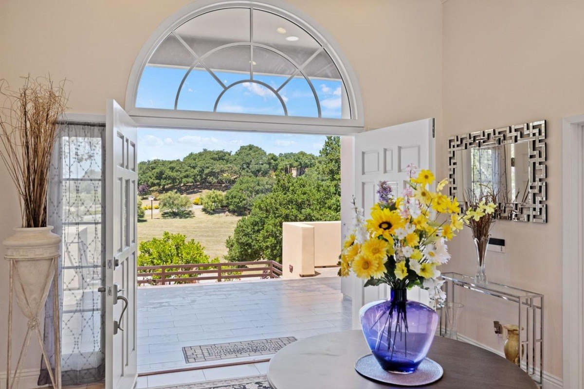 Grand entranceway with an arched window framing a scenic vista, a circular table adorned with a vibrant floral arrangement, and a mirrored console table.