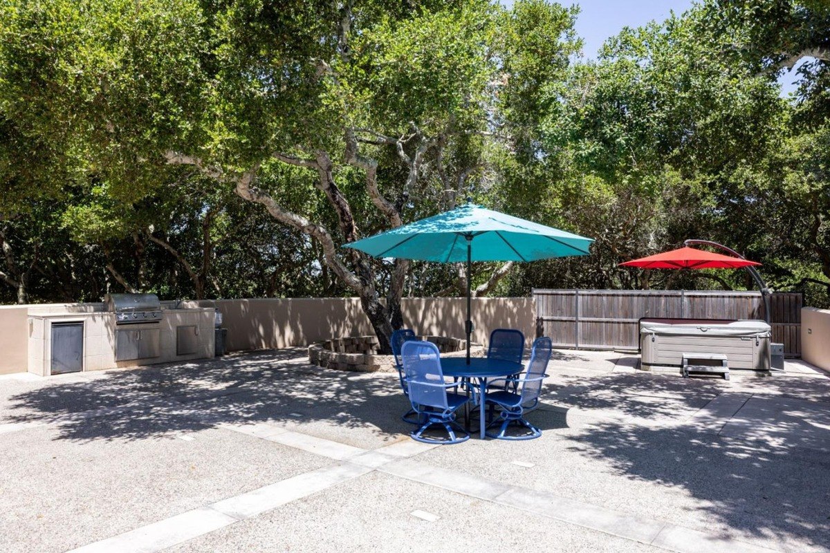 Well-appointed outdoor entertaining space featuring a built-in barbecue, a hot tub, a seating area with a table and umbrella, and lush greenery providing shade.