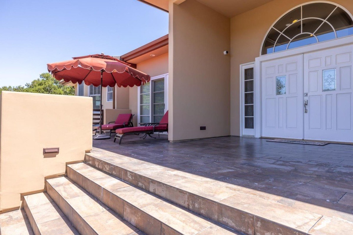 Welcoming entrance featuring a tiled patio with lounge chairs, a red umbrella, and a set of steps leading to a double-door entryway.