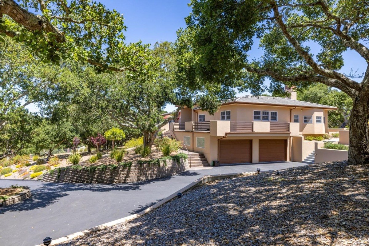 Residence with a warm stucco exterior, a terracotta roof, and a two-car garage, nestled amidst a lush landscape with mature trees.