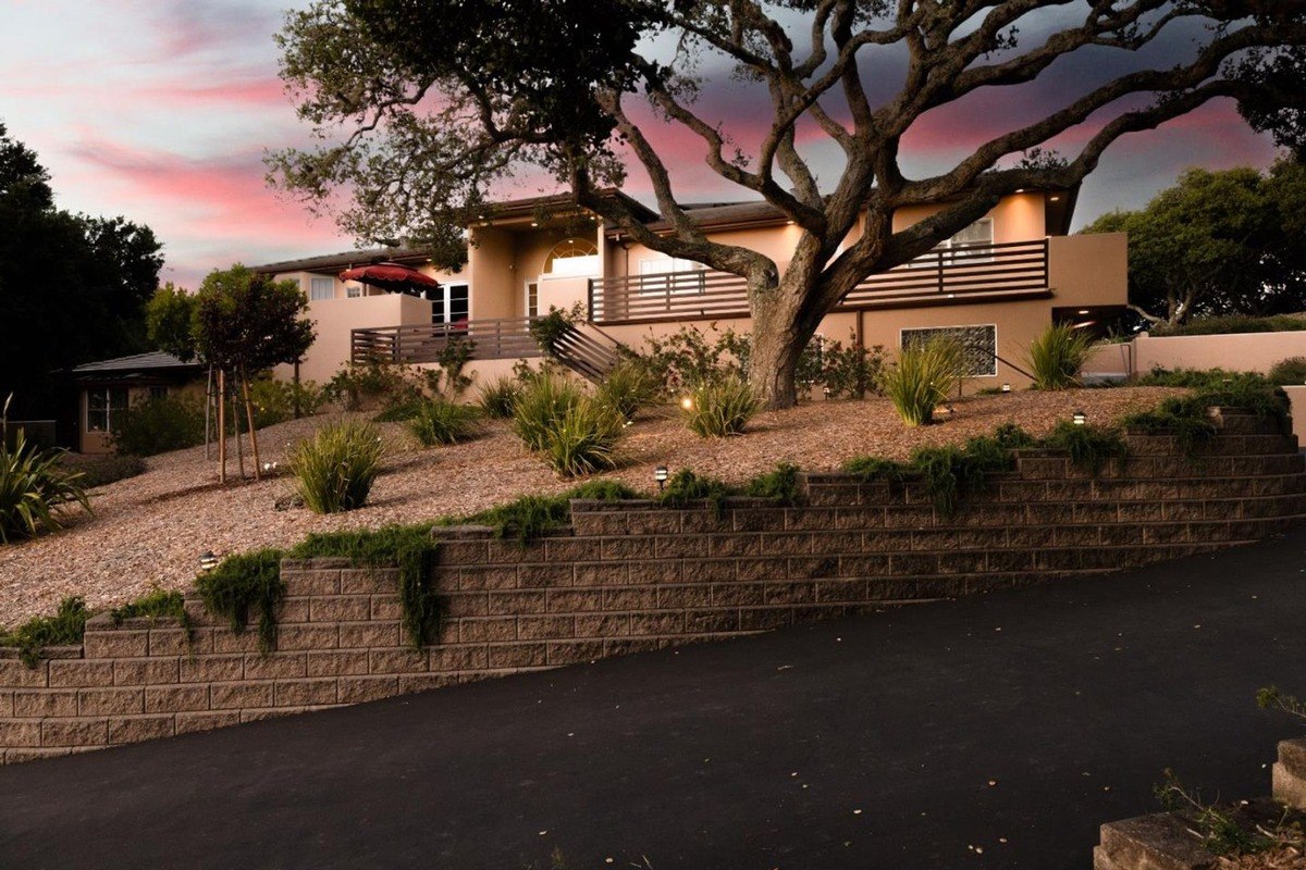 Contemporary residence with a warm stucco exterior and a landscaped front yard featuring mature trees, retaining walls, and a paved driveway, bathed in the soft glow of the setting sun.