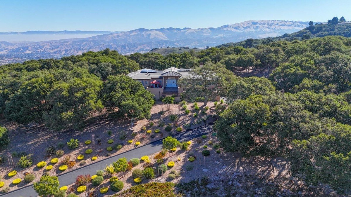 Aerial view of a secluded estate nestled amongst mature trees, showcasing a contemporary residence with a wrap-around balcony and a sweeping driveway, offering breathtaking panoramic views of the surrounding mountains.