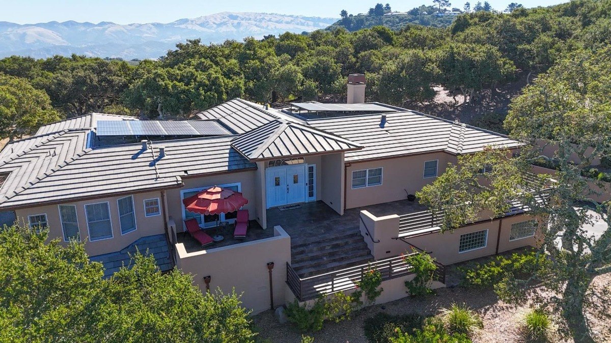 Aerial view of a contemporary residence showcasing a metal roof, solar panels, and a multi-level design, nestled amongst a picturesque landscape with rolling hills in the distance.