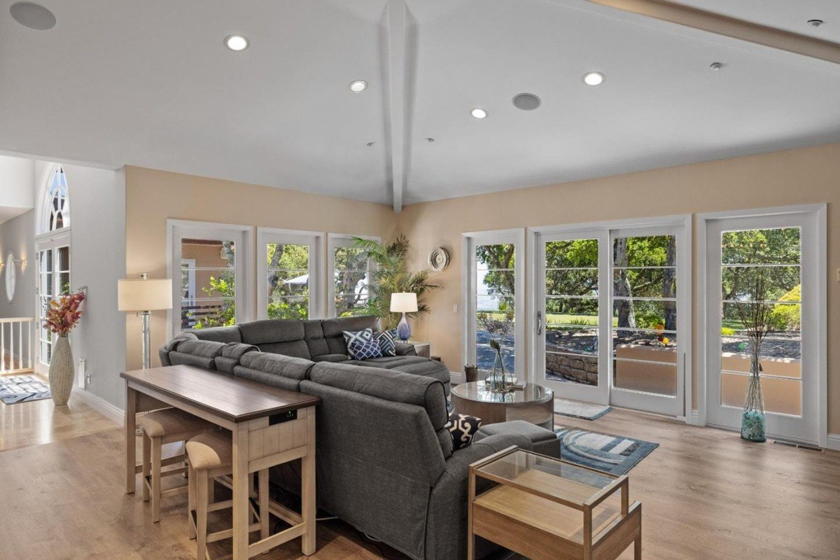 Living room with a high ceiling and lots of windows. There's a long couch and some chairs.