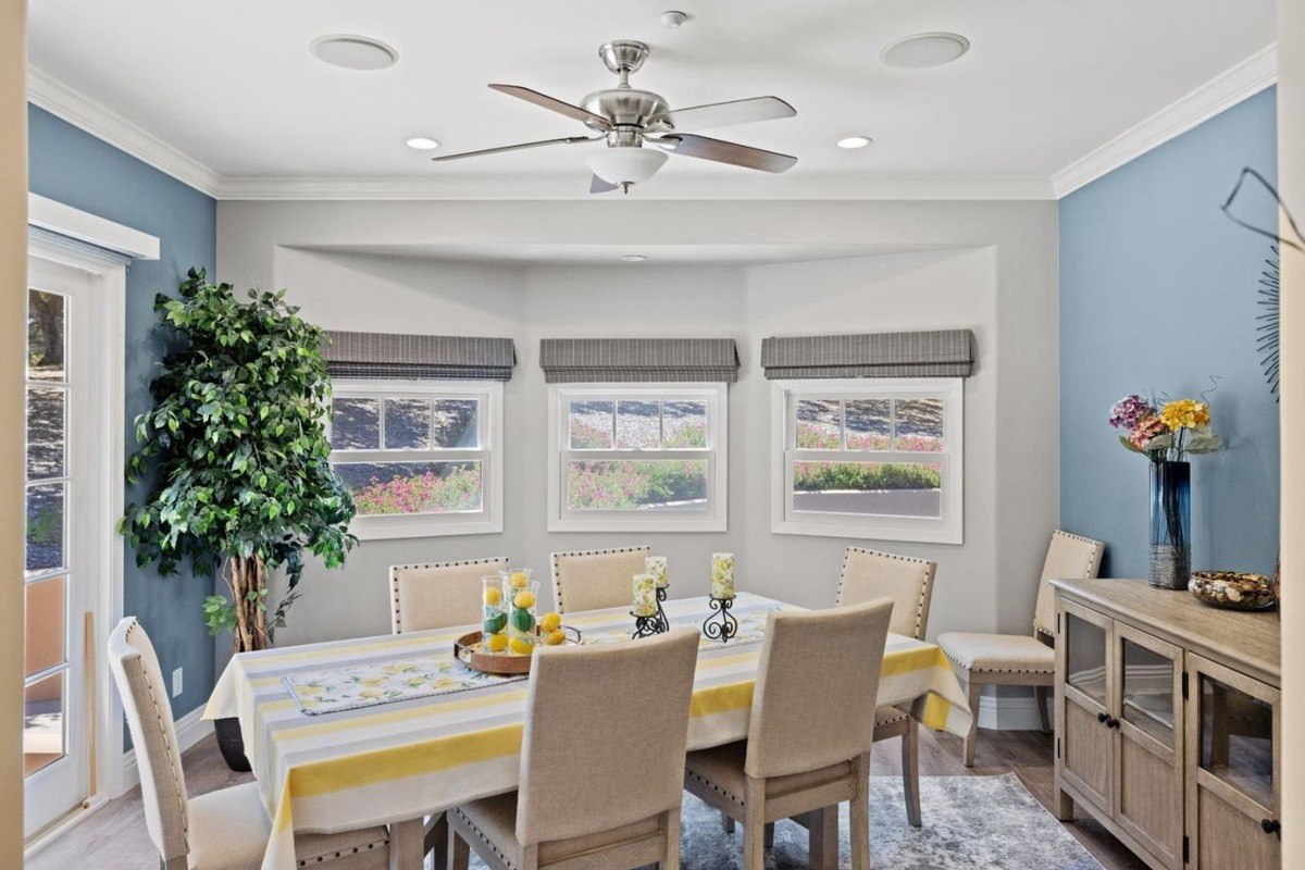 Sunlit dining room with a bay window, featuring a table set for a meal, a ceiling fan, and a vibrant accent wall.