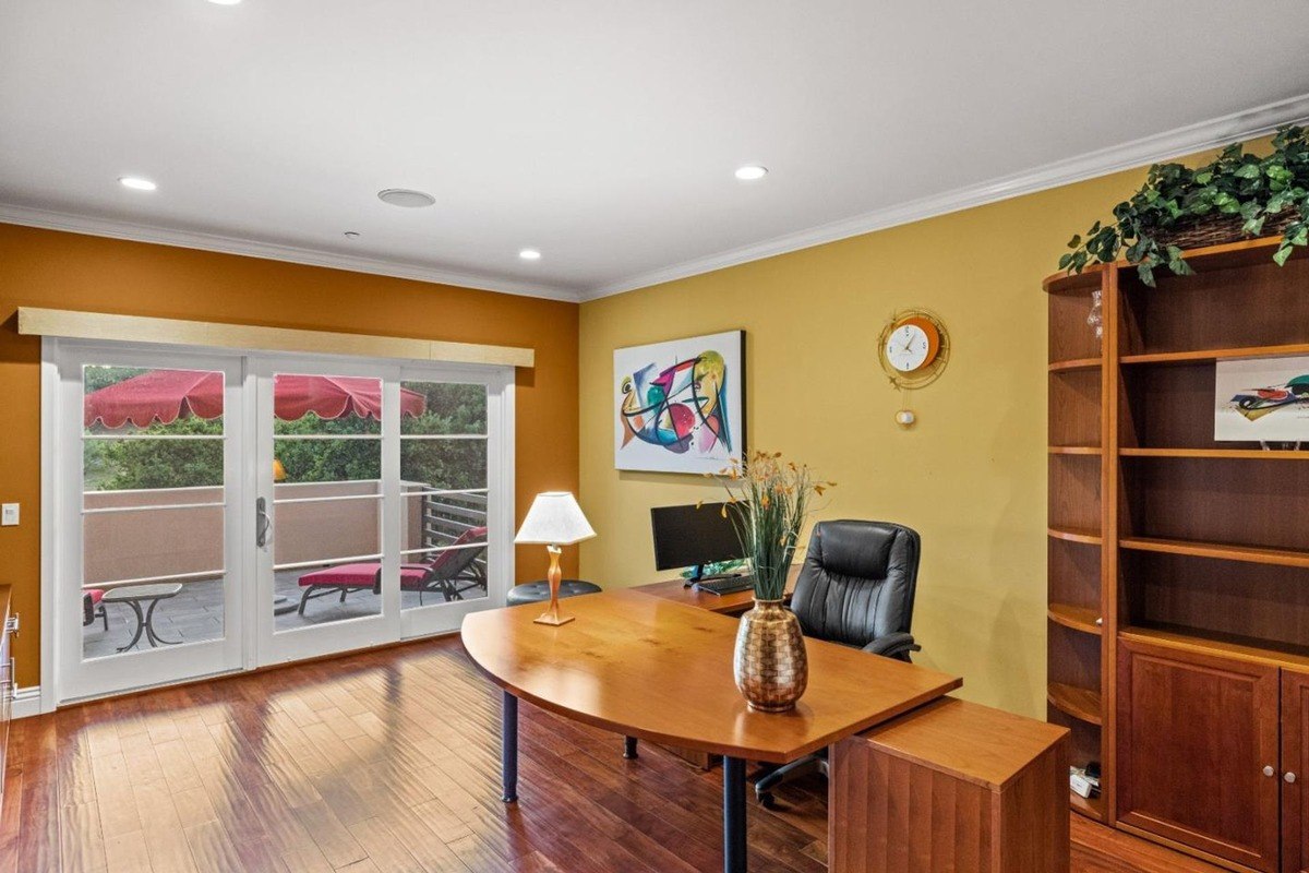 Home office with a large desk, a comfortable chair, and built-in bookshelves, offering a view of a private patio through French doors.