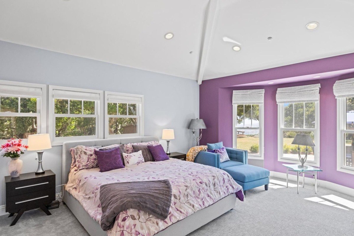 Spacious bedroom with a vaulted ceiling, featuring a king-sized bed adorned with plush bedding, a chaise lounge, and large windows offering a picturesque view.