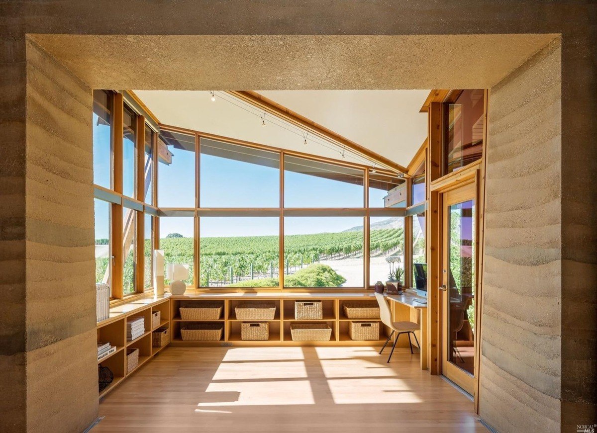 Room has rammed earth walls, wood floors, large windows overlooking vineyard, and built-in shelving.