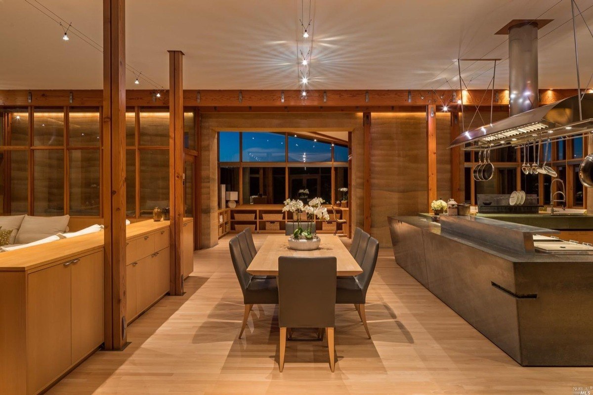 Dining space and open kitchen illuminated by soft evening lighting with sleek furnishings and polished wood beams.