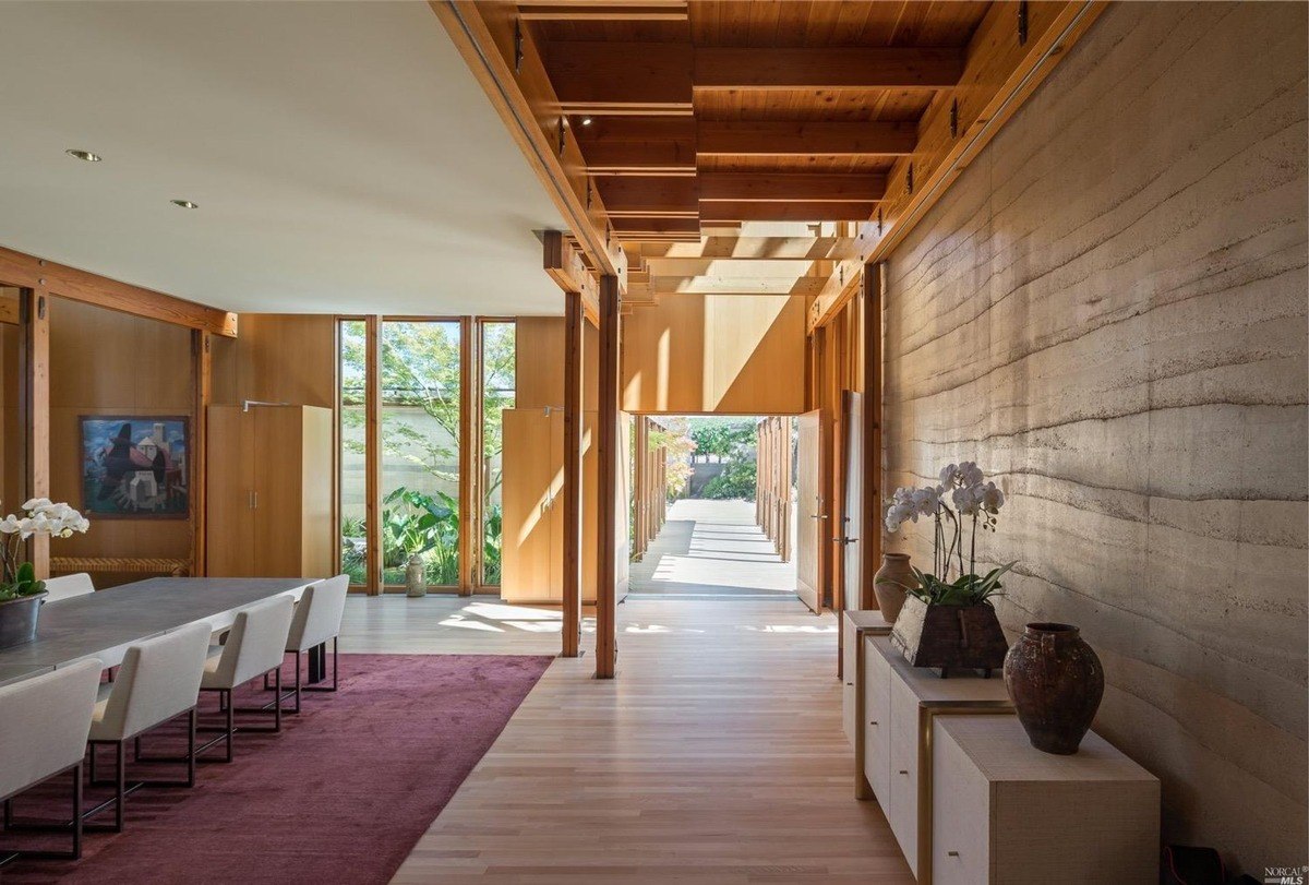 Dining space with wood beams, tall windows, and stone-textured walls, illuminated by natural light.