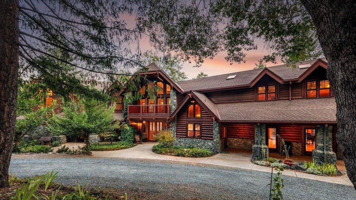 Log-style home framed by trees features a long driveway with warm exterior lighting and multiple large windows.