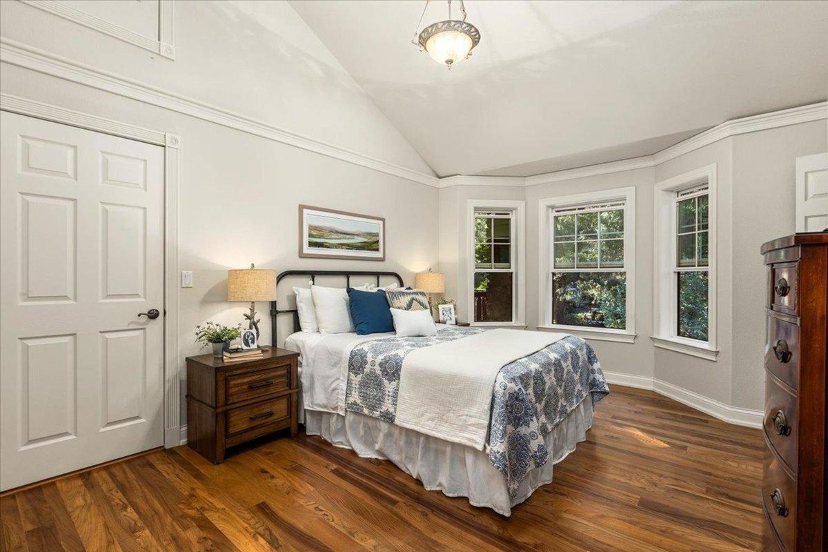 Bedroom featuring a vaulted ceiling, neutral decor, and bay windows that overlook lush greenery.