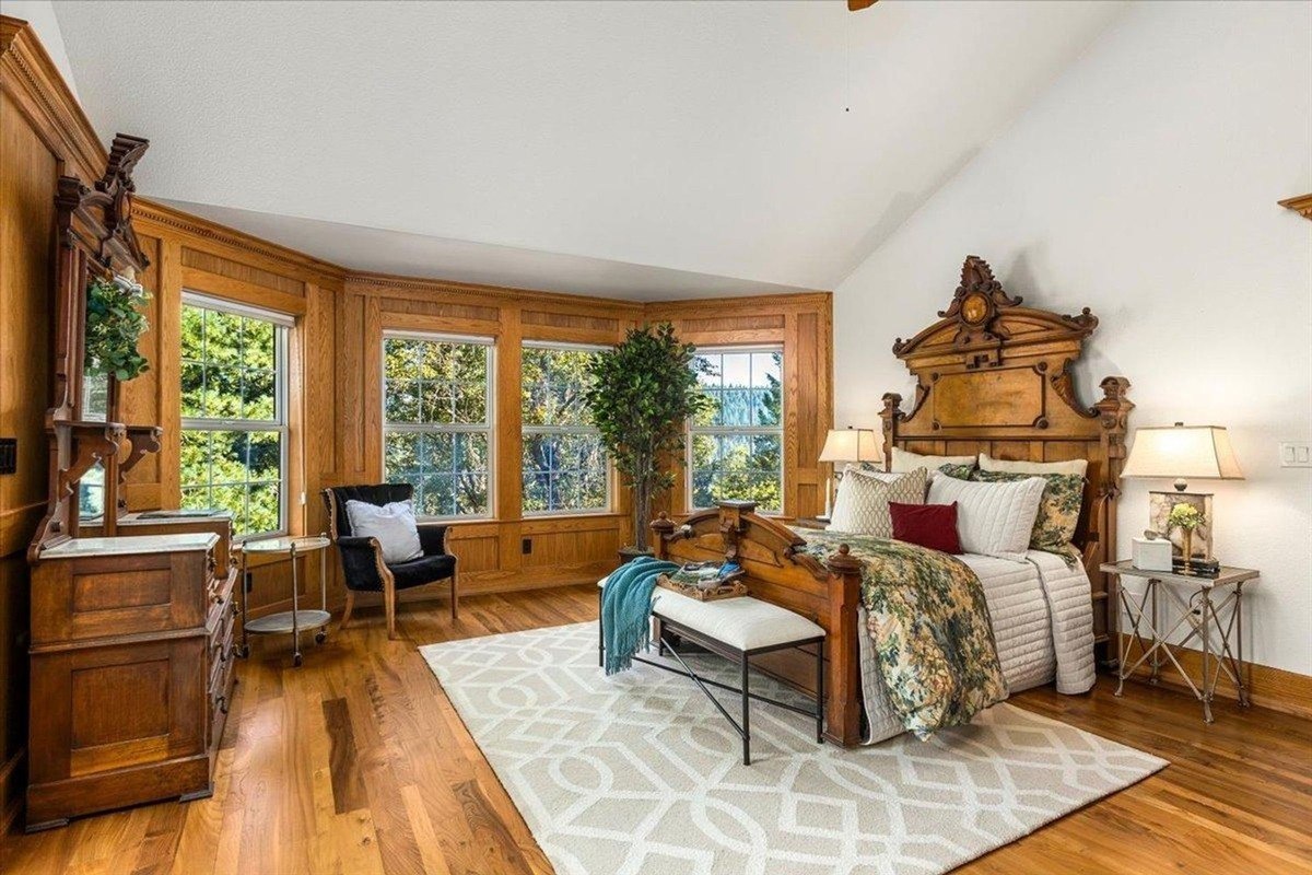 Elegant primary suite with ornate wooden furniture, expansive bay windows, and natural wood flooring.