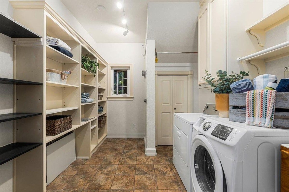 Laundry room features built-in shelves, a washer, and a dryer with warm beige cabinetry and tiled flooring.