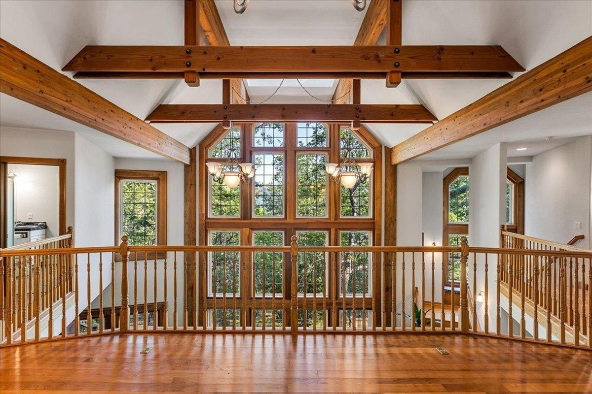 Upper-level landing provides a sweeping view of large windows and exposed ceiling beams, enhancing natural light and architectural appeal.