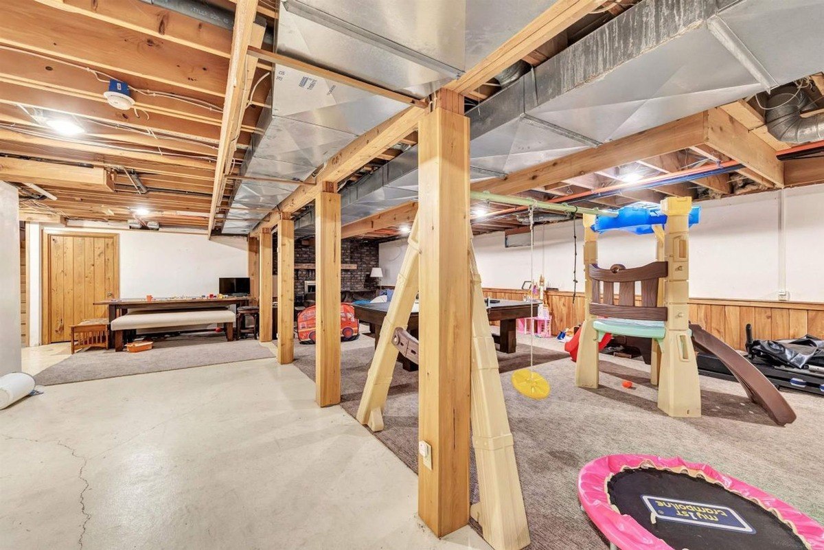 Large basement play area features exposed wood beams, support posts, and a mix of carpeted and concrete sections.