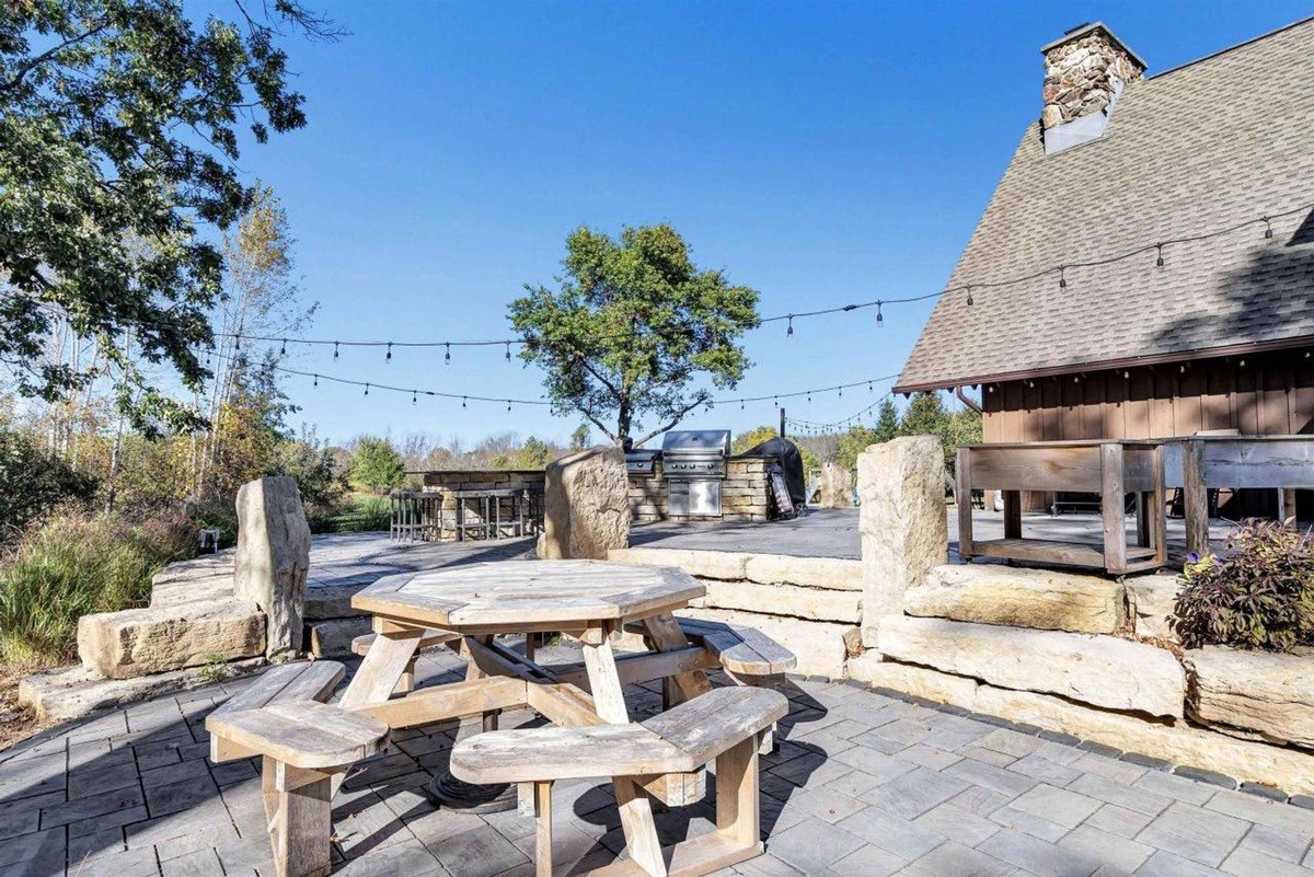 Outdoor seating area highlights a rustic wooden picnic table, built-in stone grilling station, and overhead string lights.