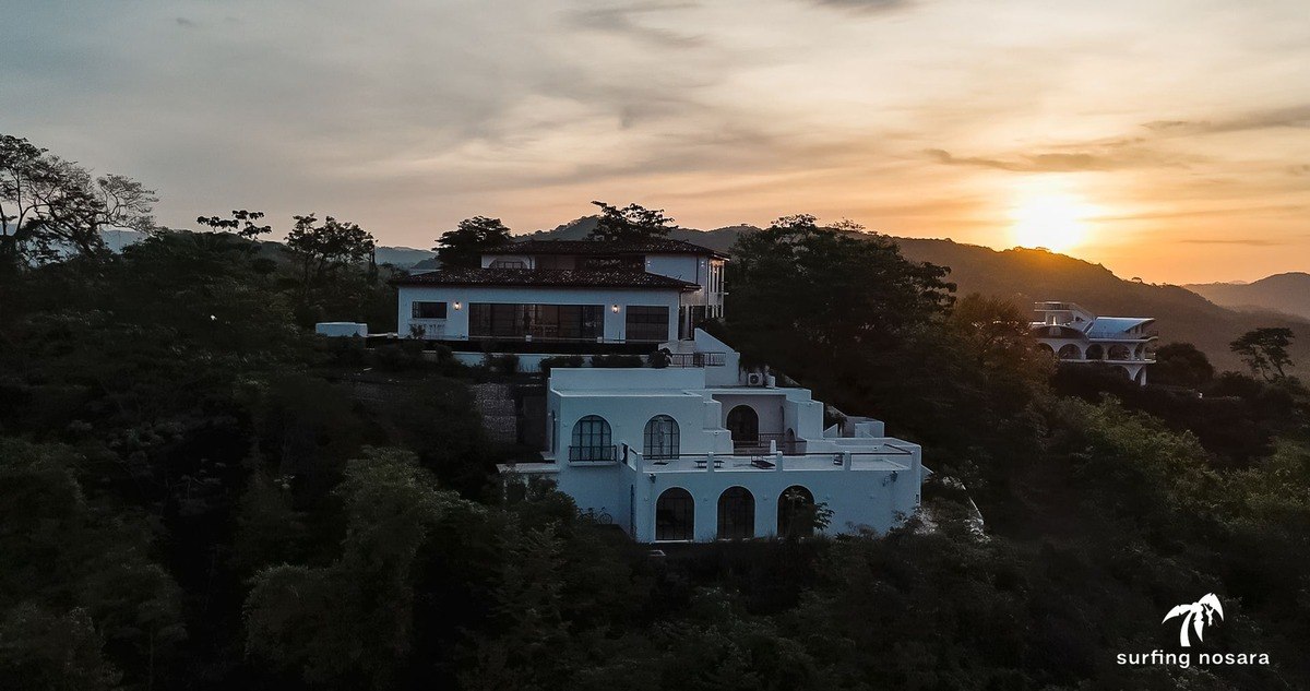 White house perched on a hillside are silhouetted against a vibrant sunset over rolling hills.