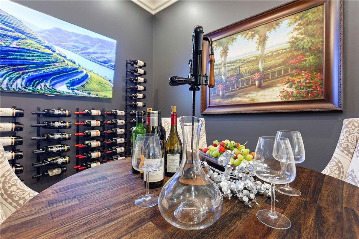 Wine tasting room features a wooden table set for wine tasting, with bottles, glasses, and fruit, plus a large wine rack and artwork on the walls.