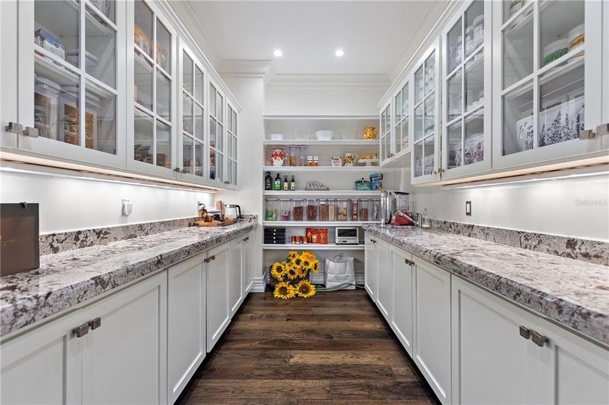 Large pantry features white cabinets with glass doors, granite countertops, and dark hardwood floors, with shelves stocked with food items and a bouquet of sunflowers.