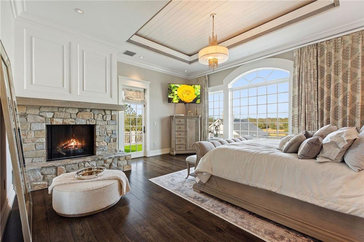 Master bedroom features a large bed, stone fireplace, hardwood floors, and large windows offering a scenic view.