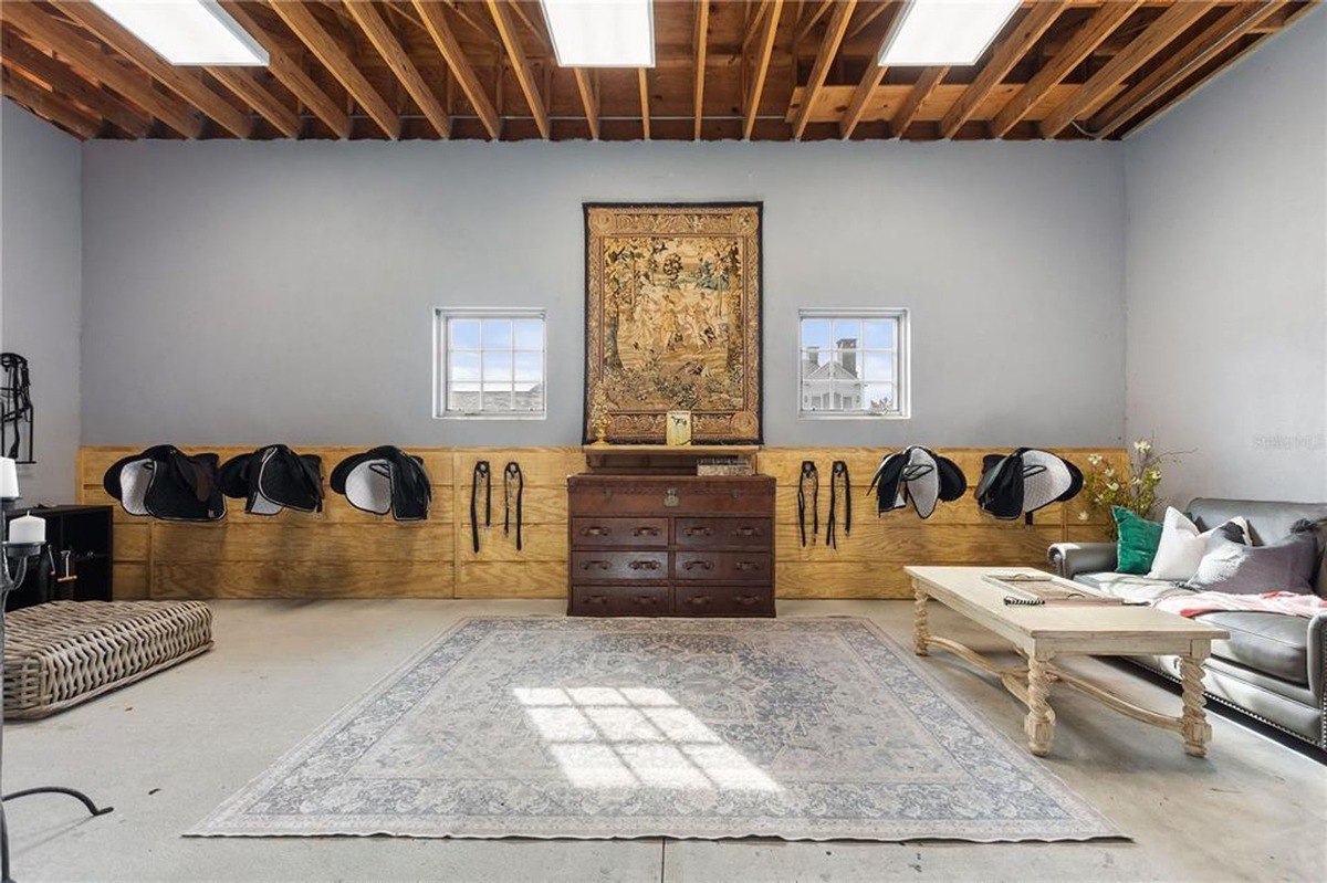 Tack room features saddles hanging on the wall, a large rug, and a wooden chest.