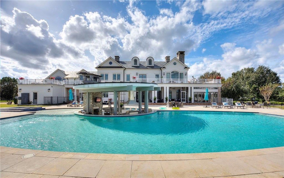 Large pool with a swim-up bar is situated in front of a large white house under a partly cloudy sky.