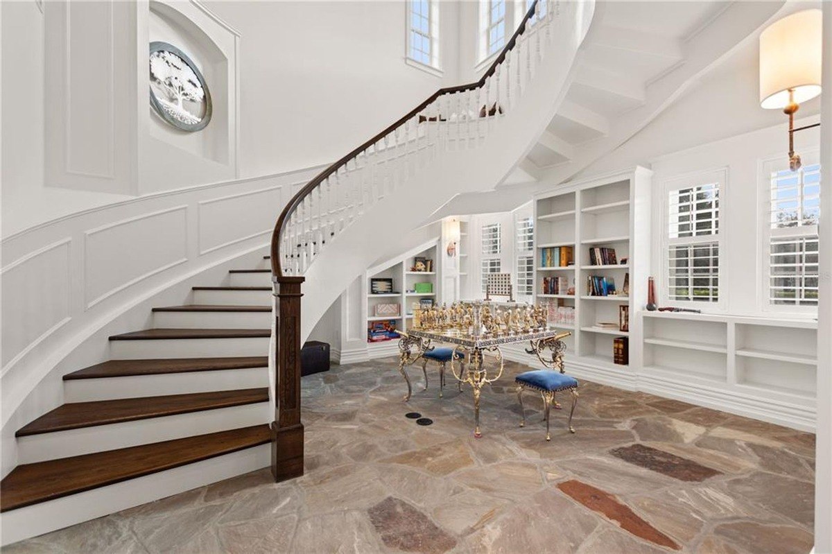 Grand foyer features a curved staircase with a dark wood railing, built-in bookshelves, and a stone floor.