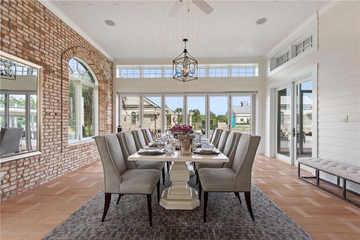 Bright dining room has a large table with gray chairs, brick walls, and floor-to-ceiling windows and doors that open to a patio.